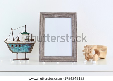 Mock up picture frame on white shelf with wooden boat and wooden airplane against white wall; portrait orientation; stylish interior mockup background with modern accessories