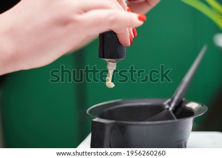 Close-up picture of hairdresser's tools in female hands. Process of squeezing hair dye from tube into plastic bowl. Hair stylist preparing for dying hair in barber shop. Beautification.