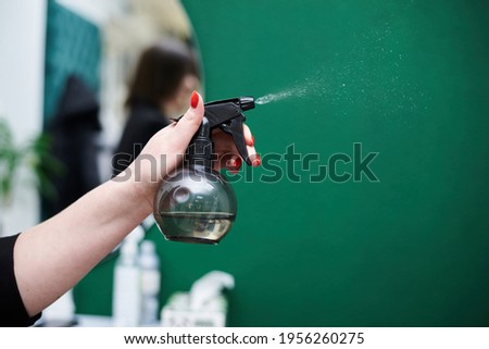 Close-up picture of hairdresser's tools in female hands isolated on green background. Small black sprayer pulverizing water for perfect haircut by hair stylist. Work process in barber shop.