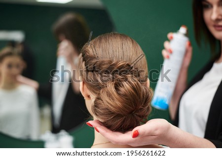 Bride getting ready for wedding. Professional hairdresser making coiffure for female client, applying hairspray in front of big mirror. Work process in beauty studio. Close-up picture of gala hairdo.