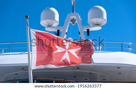 Maltese old flag white cross on red background waving on yacht stern. Luxury boat anchored at marina in Greece. Blue sky background, close up view.