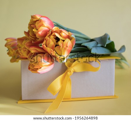 Orange-red fresh tulips in a bouquet and a gift box on a yellow background. Greetings, celebration, romance concept. Copy space for your text. The postcard for congratulations on holidays and events