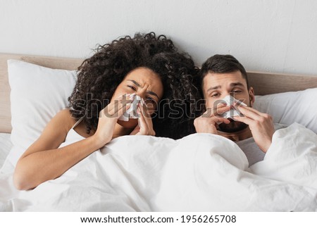 sick interracial couple holding napkins and sneezing while looking at camera