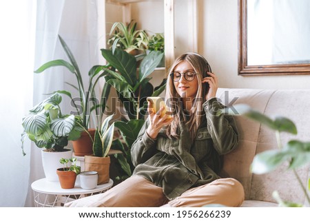 Freelance woman in glasses with mobile phone listening music in headphones and relax at home. Happy girl with closed eye sitting on couch in living room with plants.  Royalty-Free Stock Photo #1956266239