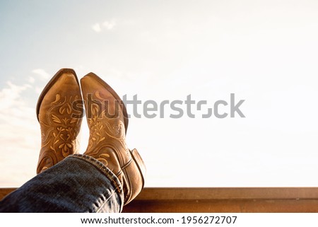 cowboy resting legs with feet crossed - sky background - negative space - boots Royalty-Free Stock Photo #1956272707