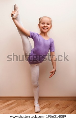 A little girl in a purple leotard raised her leg. The little ballerina trains at home. The child does warm-up and stretching