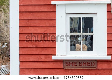 A small closed vintage glass window with wide white trim. The exterior wall of the building is a bright red narrow wood clapboard. There's a sign with the words she shed under the window ledge.