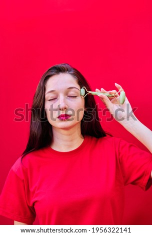 Vertical photo of a young brunette girl giving herself a facial massage using a jade roller on flat red background