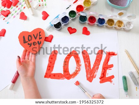 Child decorated with glitter word love. making homemade greeting card. A little girl paints a hearts as a gift for Mothers Day or Valentines day. Traditional play concept. Arts and crafts concept.