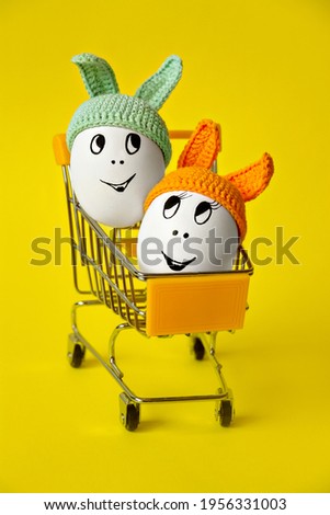 two white happy eggs with cheerful faces in green and orange beanie with rabbit ears in shopping cart on yellow background