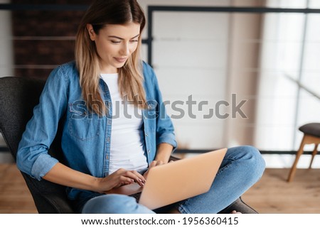 Focused lovely woman smiling and looking at laptop screen while sitting cross-legged in arm-chair