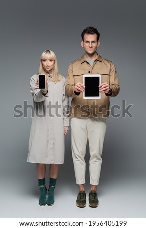 full length of trendy models holding gadgets with blank screen on grey