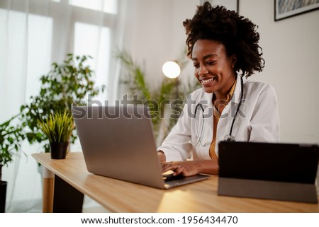 Successful young Afro American female doctor in white medical uniform and stethoscope busy working on laptop in modern hospital, smiling woman nurse or GP typing on computer consult patient online. Royalty-Free Stock Photo #1956434470