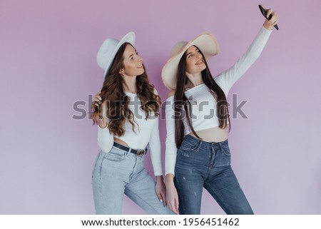 Image of two young happy women friends standing over pink wall. Looking aside make selfie by phone.