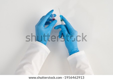 Hands of a health worker in blue gloves on a white background hold a glass ampoule with a red medicine and a syringe for injections for vaccines