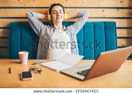 Positive brunette woman with yellow headphones listens to music having break at wooden table with modern laptop in spacious light coworking office. 
