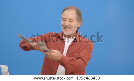 Old man with beard in front of a blue background who is cheerfully gesturing while handing out money. Concept of to pour money without thinking. It makes it rain by throwing money into the air. 