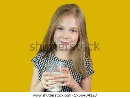 A cute little girl with blonde hair holds and drinks milk from a glass. It stands on a yellow background.Milk is good for children. She is healthy and happy. There is a place for the text.Soft focus.