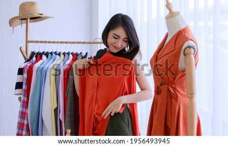 Smiling Asian woman Choose and try on clothe orange color in the Tailor shop. Fashion designer standing in clothing In order to repair, service for customers. Concept Profession Dressmaker designer Royalty-Free Stock Photo #1956493945
