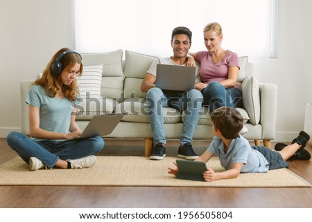 Stay at home caucasian parents working on laptops while sit on sofa and son lying on the floor watching cartoon on tablet and teenage daughter studying with laptop doing online research homework.