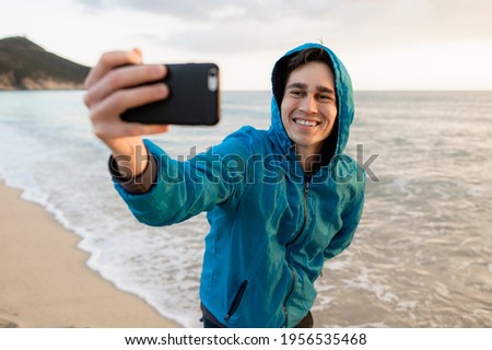 Young happy male doing a selfie with mobile phone on the beach in Sardinia during sunset light.