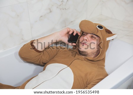 Man in cosplay costume of a cow. Guy in the funny animal pyjamas lie in the bath and speak on the mobile phone.
