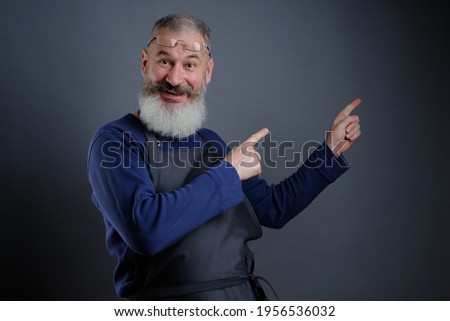 Follow me. Look here. Close up portrait of cheerful pleased qualified experienced joyful apron-wearing grandfather pointing on empty blank place over shoulder isolated on gray background