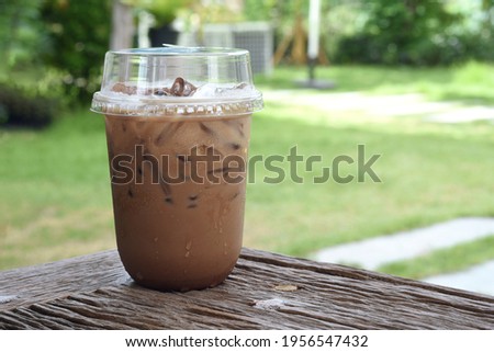 Front View of Iced Mocha Coffee in transparent plastic glass on wooden table's corner, selective focus on glass with blurred background of garden
