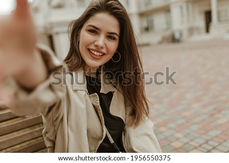 Brown-eyed attractive brunette woman in good mood looks into camera and takes selfie. Pretty lady in trench coat sits on wooden bench outside.