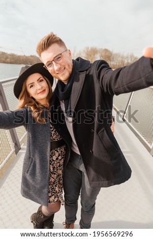 Young couple is walking outside by the lake and taking a selfie. Spend time together outside. Young people dressed in boho style. Influencers take a picture of them kissing 