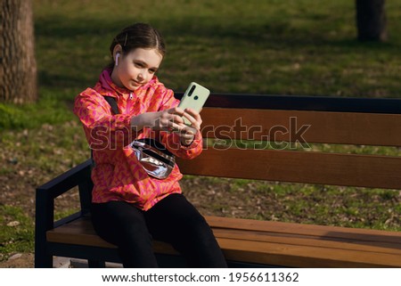 A girl sits on a park bench and takes pictures with her smartphone. The girl is stylishly dressed and listens to music with headphones using modern technologies. Free space for text