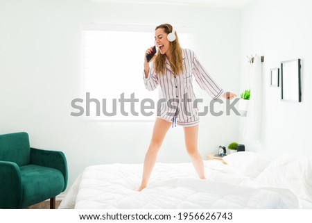 Latin woman with headphones playing karaoke and singing a song while dancing in her bed. Beautiful woman in pajamas having fun in her bedroom