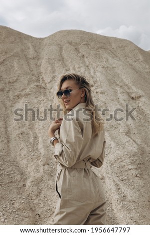 A slender woman stands in the middle of the desert. The blondes eyes are hidden behind sunglasses. A persons clothes merge with the surrounding environment. High quality photo