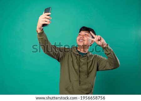 Smiling adorable male making selfie photo on smartphone with positive expression in casual clothing and looking at camera over green background. Happy adorable glad man rejoices success.