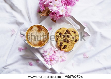 Big cup of coffee with milk and chocolate cookies on the white bedsheet. Sakura flowers around cut of hot coffee and dessert. Top view. Spring time. Wooden plate with coffee and flowers.
