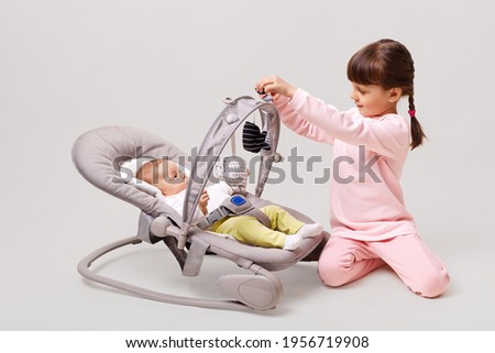 Baby girl lying down in cradle with her sister, little girl with pigtails sitting on floor near her newborn sis, posing isolated over white background. Royalty-Free Stock Photo #1956719908