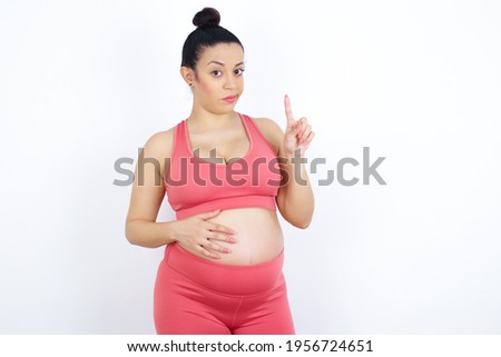 No sign gesture. Closeup portrait unhappy young beautiful Arab pregnant woman in sports clothes against white wall raising fore finger up saying no. Negative emotions facial expressions, feelings.