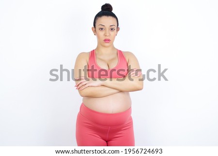 Picture of angry young beautiful Arab pregnant woman in sports clothes against white wall looking camera.