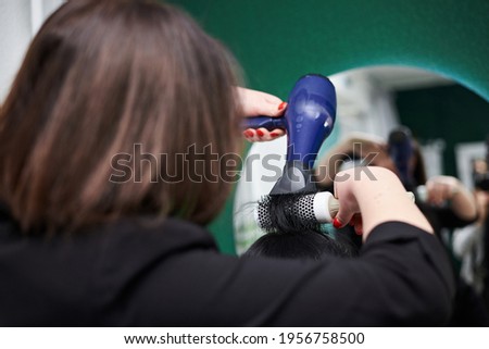 Young brunette woman getting ready for party. Professional hairdresser making hairstyling for female client with hair dryer in front of big mirror. Close-up picture of work process in barber shop.