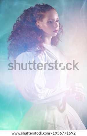 Portrait of a refined fashion model girl with lush red curly hair posing in a long white haute couture dress among flashes and haze on green background. Studio shot. Art and fashion.  