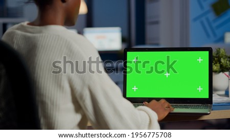 African woman manager looking at mock up monitor of laptop, green screen mockup, chroma key desktop sitting at desk in business office late at night. Freelancer watching at isolated desktop overwork
