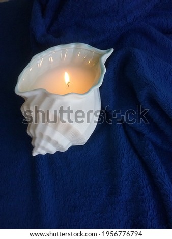 White sea shell candle, lit with warm glowing flame, set in a long portrait angle against a scrunched navy blue polar fleece blanket. 