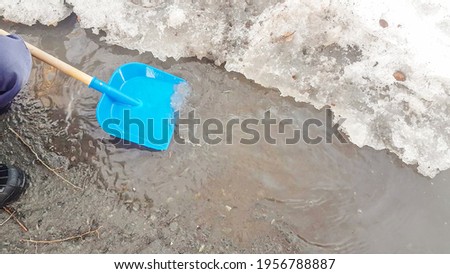 Close-up of a child's feet in rubber boots, a boy child happily playing with a shovel in a muddy puddle with snow and ice, in the early spring in the yard.