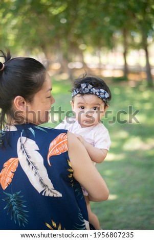 Smiling Asian mother holding a cute little baby girl on blured background of green garden, baby looking at camera 