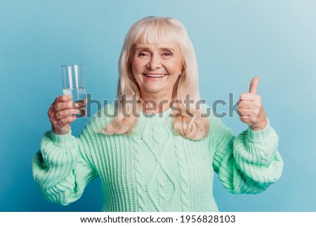 Elderly lady hold glass of pure mineral water show thumb-up gesture isolated on blue background
