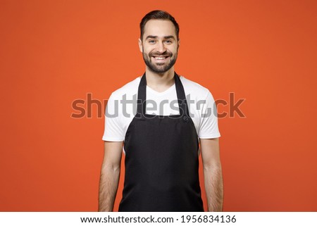 Young fun man 20s barista bartender barman employee in black apron white t-shirt work in coffee shop looking camera isolated on orange color background studio portrait. Small business startup concept Royalty-Free Stock Photo #1956834136