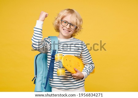 Joyful little male kid teen boy 10s years old in striped sweatshirt eyeglasses backpack hold skateboard doing winner gesture isolated on yellow background, child studio portrait. Education concept