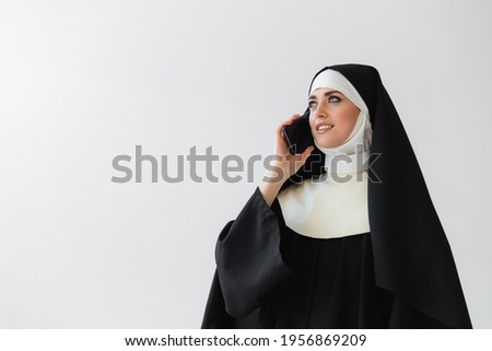joyful nun in black mantle talking on mobile phone isolated on grey