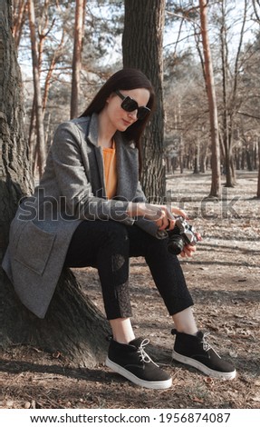 A girl in black glasses and a gray coat sits near a large tree in the forest and looks at the camera