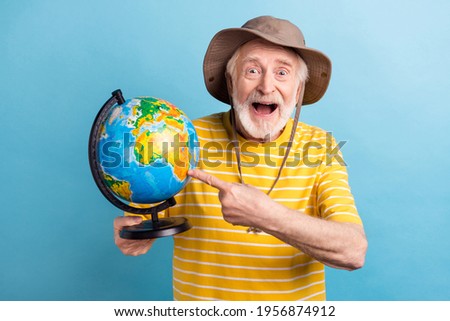 Photo of happy positive amazed grey haired old man point finger globe travel wear hat isolated on pastel blue color background